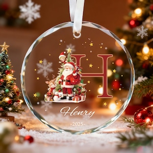 May include: A circular glass Christmas ornament with a faceted edge, featuring a printed illustration of Santa Claus, a reindeer, and an elf. The design includes a large letter H, the name Henry, and the year 2025, suspended by a white ribbon.