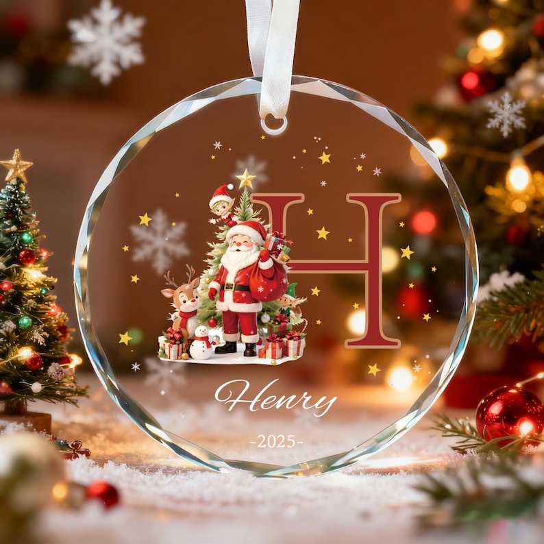 May include: A circular glass Christmas ornament with a faceted edge, featuring a printed illustration of Santa Claus, a reindeer, and an elf. The design includes a large letter H, the name Henry, and the year 2025, suspended by a white ribbon.
