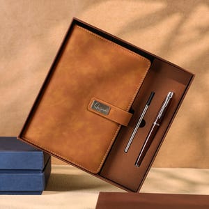 Op de afbeelding: Een bruin leren notitieboek en pennenset in een geschenkdoos. Het notitieboek heeft een sluiting met een metalen naamplaatje. Een zilveren pen en een donkerrode pen zijn inbegrepen. De doos is bruin en het notitieboek heeft een lichtere bruintint.