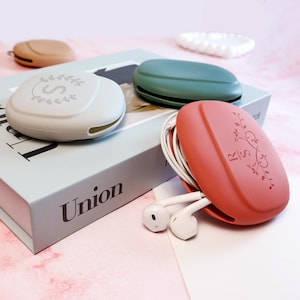 Op de afbeelding: Een verzameling kleurrijke, ovale oortelefoonhoesjes. E&eacute;n is koraal met een bloemenontwerp en oortelefoons, een andere is saliegroen en een derde is lichtgrijs met een "S" en een kransontwerp. De hoesjes liggen op een boek.