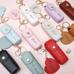 Puede incluir: Una colección de llaveros personalizados en varios colores, incluyendo negro, rojo, rosa y azul. Cada llavero presenta una funda rectangular con cierre de botón a presión y un llavero dorado. Algunos llaveros tienen nombres grabados y otros diseños florales.