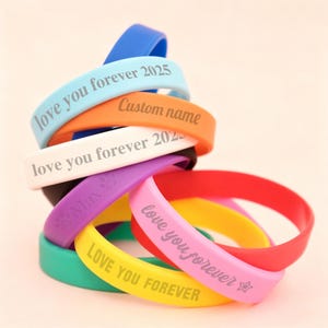 May include: A stack of colourful silicone wristbands in blue, light blue, orange, white, black, purple, red, yellow, and teal. The bands feature engraved text including "love you forever 2025", "Custom name", "Max" with a paw print, and "LOVE YOU FOREVER".