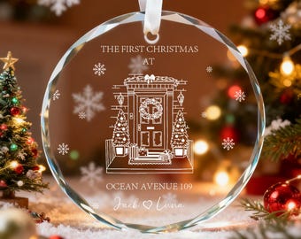 May include: A clear, faceted glass Christmas ornament with a white ribbon. The ornament features an etched design of a house with a wreath, Christmas trees, and snowflakes. Text includes "THE FIRST CHRISTMAS AT OCEAN AVENUE 109" and names.