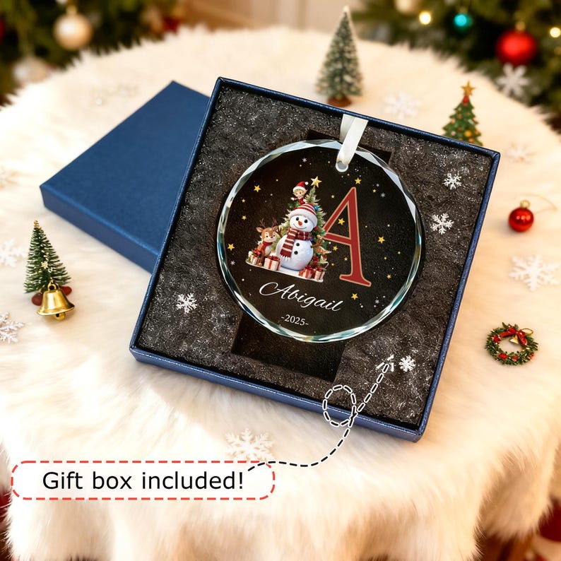 May include: A round glass Christmas ornament featuring a snowman, a reindeer, and a Christmas tree graphic with the letter A, the name Abigail, and the year 2025. The ornament is presented in a dark blue gift box with black foam padding.