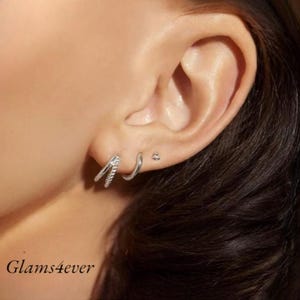 Peut inclure: Ensemble de boucles d'oreilles argentées. L'ensemble comprend une boucle d'oreille double avec une rangée de petites pierres, une boucle d'oreille simple et une petite boucle d'oreille à clous. Les boucles d'oreilles sont présentées sur une oreille. Le texte "glams4ever" est en bas à gauche.