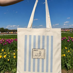 May include: Cream-colored tote bag with light blue vertical stripes. A postage stamp-style graphic in the center features a shepherd and sheep. The bag is held up against a blue sky and a field of flowers.
