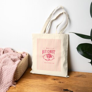 May include: A cream-colored canvas tote bag featuring a vertical pink striped rectangular design. Centered is a dark pink graphic of a rose with the text "NOT I BUT CHRIST IN ME" and "GALATIANS 2:20" printed in a decorative serif font.