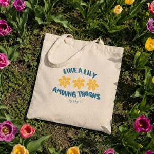May include: A cream-colored tote bag with the text "LIKE A LILY AMONG THORNS" in blue and yellow. The bag is surrounded by colorful tulips in a garden setting. The bag has two handles.