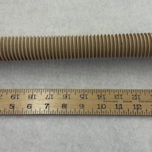 May include: A light beige threaded bolt with a hexagonal nut on each end, positioned above a wooden ruler. The bolt measures approximately 8.5 inches in length, with the ruler showing the scale from the 1-inch mark to the 9.5-inch mark.