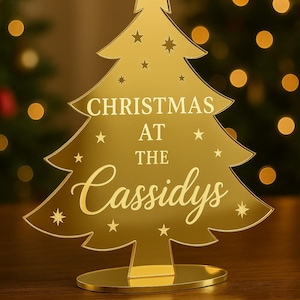 Może przedstawiać: Złota dekoracja choinkowa z napisem "CHRISTMAS AT THE Cassidys". Choinka ma gwiazdę na górze i gwiazdki. Ozdoba stoi na okrągłej podstawie.