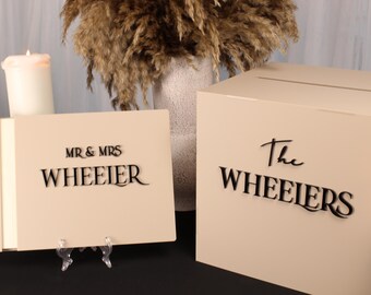 May include: Beige wedding card box and guest book with black lettering that reads "MR & MRS WHEEIER" and "The WHEEIERS". The guest book is propped up on a clear stand. A lit candle and decorative dried flowers are in the background.
