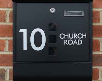 May include: A black mailbox mounted on a red brick wall. The mailbox has the number "10" and the words "CHURCH ROAD" in white lettering. It features a keyhole and a small window.
