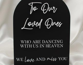 May include: A black arched sign with a rectangular base. The sign has white script that reads "To Our Loved Ones, Who Are Dancing With Us In Heaven, We Love and Miss You."