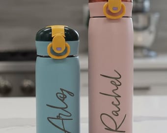 May include: Two insulated water bottles, one teal and one pink, with yellow accents on the lids. The teal bottle has the name "Arlos" in script, and the pink bottle has "Rachel" in script. Both bottles are on a white surface.