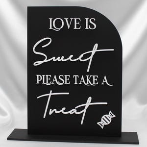 May include: Black sign with white text that reads "LOVE IS Sweet PLEASE TAKE A Treat" with a candy graphic. The sign is on a black base and has a curved top. The background is a white, silky fabric.