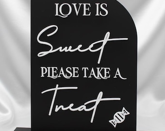 May include: Black sign with white text that reads "LOVE IS Sweet PLEASE TAKE A Treat" with a candy graphic. The sign is on a black base and has a curved top. The background is a white, silky fabric.