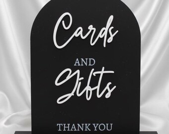 May include: A black wooden sign with an arched top, displaying the words "Cards and Gifts" in white script. The words "THANK YOU" are at the bottom. The sign is on a black base, set against a white satin backdrop.