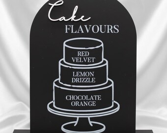 May include: Black arched sign with the words "Cake Flavours" and a tiered cake graphic in white. The cake graphic lists the flavors: Red Velvet, Lemon Drizzle, and Chocolate Orange. The sign is on a black base.
