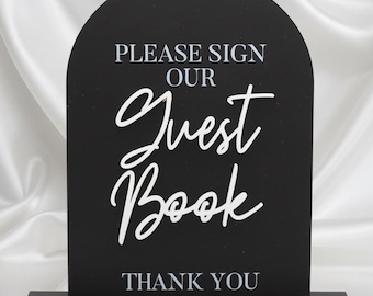 May include: Black arched sign with white text that reads "PLEASE SIGN OUR Guest Book THANK YOU". The sign is on a black base and set against a white satin background. Ideal for weddings or events.
