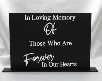 May include: A black memorial sign with white text that reads "In Loving Memory Of Those Who Are Forever In Our Hearts." The sign is rectangular and stands on a small base, set against a white satin background.