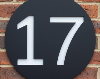 May include: A black circular house number sign with the number 17 in white. The sign is mounted on a red brick wall. The number is in a bold, sans-serif font.