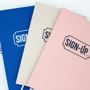 Può includere: Una collezione di quaderni bianchi, blu, beige e rosa. Ogni quaderno presenta la scritta "SIGN-UP" stampata in un design retrò blu scuro. I quaderni sono disposti a ventaglio, con le copertine visibili.