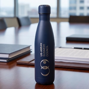 May include: A matte navy blue metal water bottle with a screw-top lid. The bottle features the white text US-ASEAN BUSINESS COUNCIL printed vertically above a circular logo consisting of two interlocking rings in a light gray or silver color.