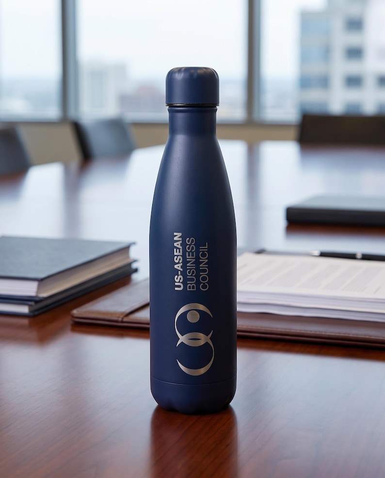 May include: A matte navy blue metal water bottle with a screw-top lid. The bottle features the white text US-ASEAN BUSINESS COUNCIL printed vertically above a circular logo consisting of two interlocking rings in a light gray or silver color.