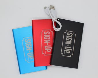 May include: Three rectangular power banks in blue, red, and black, each with the text "SIGN-UP" in a white rectangular frame. A white USB cable is connected to the black power bank. The power banks are arranged on a white surface.