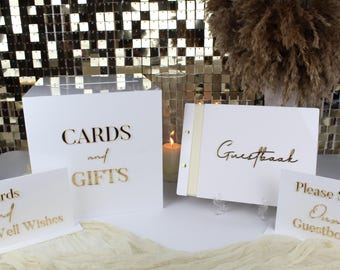 May include: White acrylic wedding decor set with gold lettering. Includes a box for cards and gifts, a guestbook, and signs that read "Cards and Well Wishes" and "Please Sign Our Guestbook."