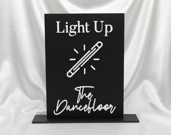 May include: Black rectangular sign with white text that reads "Light Up The Dancefloor". The sign features a glowing lightbulb graphic. The sign is mounted on a black base, set against a white satin backdrop.