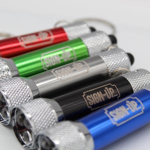 May include: A collection of small flashlights in red, green, silver, black, and blue. Each flashlight has a silver textured head and the text "SIGN-UP" printed on the body. The flashlights are attached to keychains.