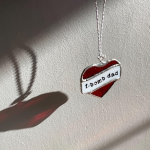 May include: A heart-shaped pendant with a red center and a white banner that reads "f-bomb dad." The pendant is outlined in silver and hangs from a delicate silver chain. The image is set against a neutral background.