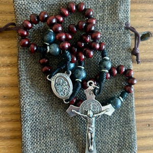 Pode incluir: Um rosário de madeira vermelha com uma medalha de prata de São José e um crucifixo de prata de São Francisco de Assis. O rosário está em uma bolsa de cordão cinza.