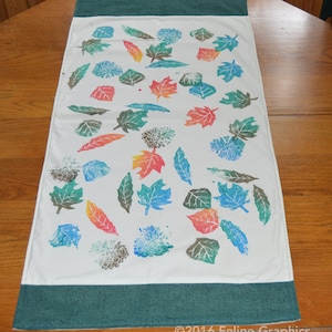 Può includere: Un runner da tavolo in tessuto bianco con un bordo verde presenta un design di stampe di foglie colorate in tonalità di verde, blu, arancione e rosso. Il design ricorda le foglie autunnali.