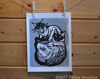 May include: Black and white linocut print of three cats, two of which are curled up together. The print is titled "The Siamese" and signed by the artist, Jen Bertram.
