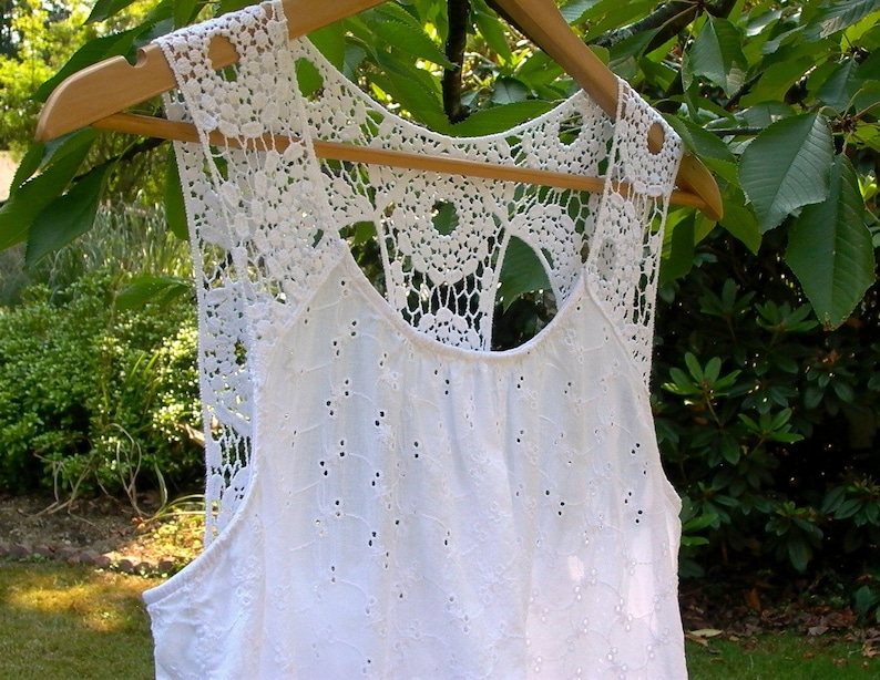 May include: White crochet top with a delicate lace design. The top has a scoop neck and a sleeveless design.