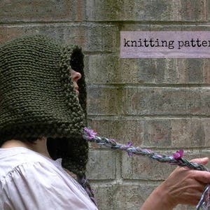 Op de afbeelding: Een donkergroene gebreide capuchon met een lange, gevlochten koord. Het koord is versierd met paarse bloemen. De capuchon wordt gedragen door een persoon die voor een bakstenen muur staat. De tekst "knitting pattern" is zichtbaar in de afbeelding.