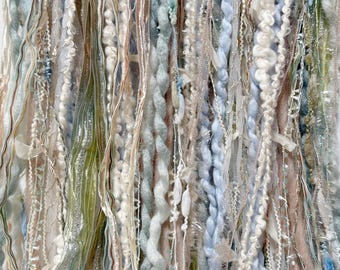 May include: Close-up of a textile art piece featuring a variety of textures and colors. The artwork incorporates strands of yarn, ribbon, and other materials in shades of blue, white, green, and beige. The piece has a layered, textured appearance.