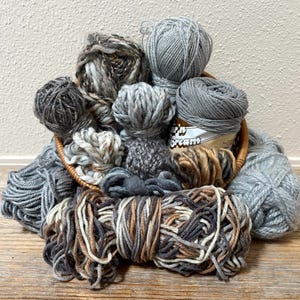 May include: A woven basket overflowing with various gray and brown yarn skeins. The yarn is in different textures and shades, including solid gray, variegated, and textured. Some yarn is loosely wound, while others are in neat balls.