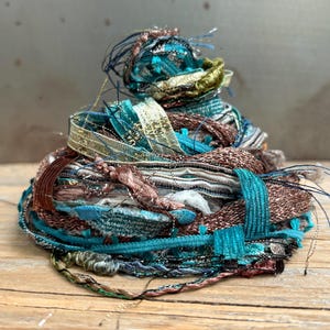 May include: A tangled bundle of assorted decorative ribbons and yarns in shades of teal, copper, brown, and metallic gold. The collection features various textures, including braided, woven, and shimmering strands layered into a dense, circular pile.