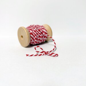 May include: A wooden spool of red and white baker's twine with a tied bow. The spool is made of light-colored wood, and the twine is tightly wound around it. The twine is red and white, creating a striped pattern.