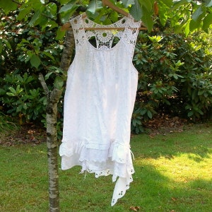 May include: White crochet dress with a lace overlay and a ruffled hem. The dress has a sleeveless design and a scoop neckline.