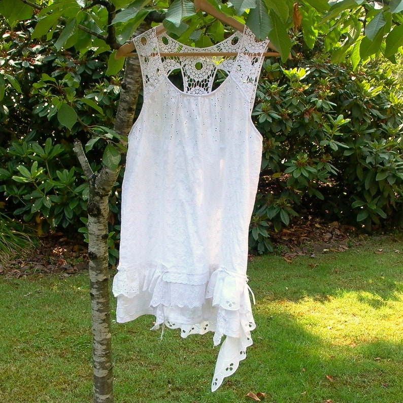 May include: White crochet dress with a lace overlay and a ruffled hem. The dress has a sleeveless design and a scoop neckline.