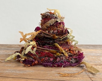 May include: A conical sculpture constructed from layered textile scraps, including ribbons, metallic threads, and lace in shades of deep burgundy, plum, copper, and gold. The textured surface features frayed edges and loose strands of fabric.