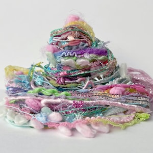 May include: A colorful pile of mixed craft supplies, including ribbons, yarn, and decorative trim. The assortment features various textures and colors, such as pink, purple, green, and silver, creating a visually appealing display for crafting projects.