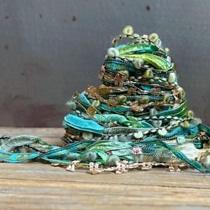 May include: A coiled bundle of decorative ribbons in various shades of emerald, teal, and mint green. The assortment includes textured velvet, metallic gold threads, frayed edges, and small fuzzy pom-pom accents layered in a dense, conical stack.