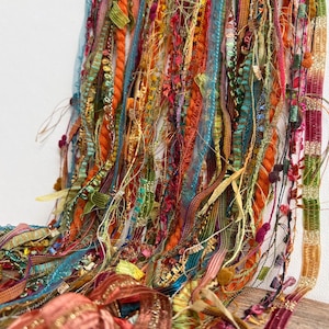 May include: A collection of hanging decorative ribbons and yarn in various textures and colors including orange, teal, green, pink, and gold. The strands feature diverse materials such as velvet, metallic thread, lace, and frayed fabric strips.