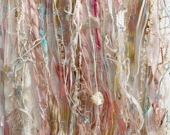 May include: A wall hanging composed of vertical strands of mixed fibers, including cream, pale pink, and soft yellow yarn, metallic silver ribbons, sheer white lace, and textured fabric strips, creating a layered, multi-dimensional textile display.