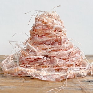 May include: A conical pile of pale pink and white decorative ribbon yarn. The yarn features a textured, open-weave construction with loose, frayed threads extending from the main structure, creating a layered, multi-dimensional appearance.